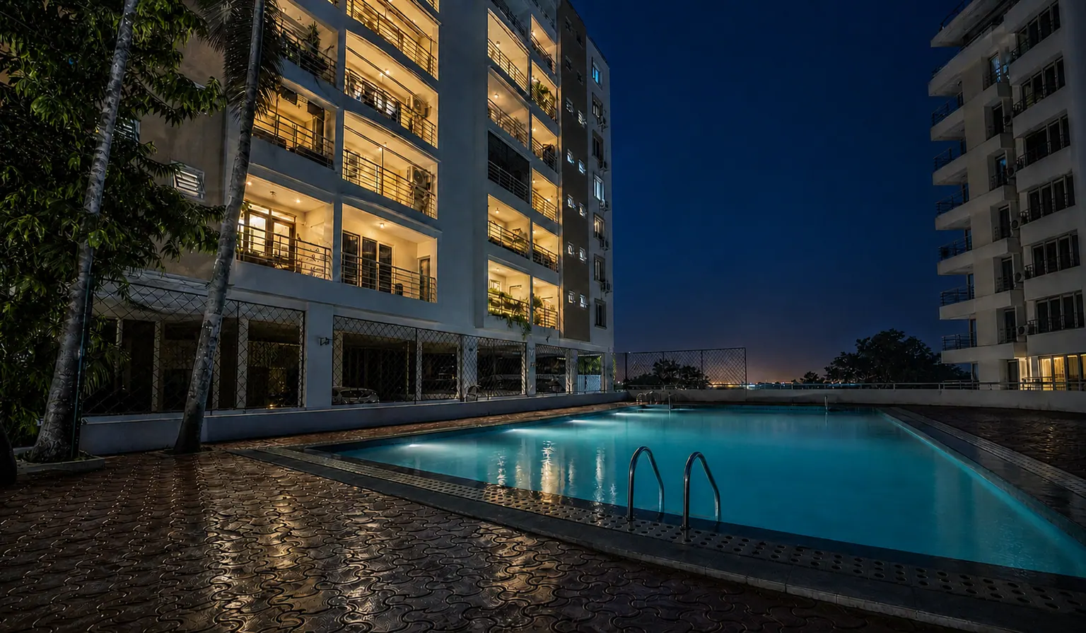 The courtyard pool lit turquoise after dark, the apartment block above it with balconies glowing warm against a deep blue sky